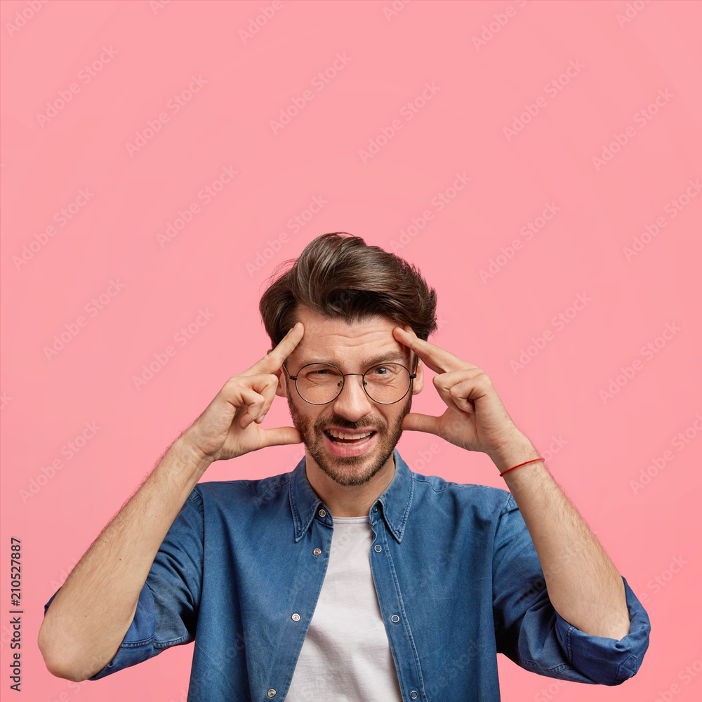 Vertical shot of stressed European male has headache after loud party, keeps hands on temples, dressed in denim shirt, has discontent expression, poses against pink background with blank space Vertical shot of stressed European male has headache after loud party, keeps hands on temples, dressed in denim shirt, has discontent expression, poses against pink background with blank space