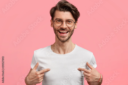 Horizontal shot of handsome cheerful hipster male indicates at white t shirt, shows blank space for your logo, being in high spirit, isolated over pink blank wall. Positive emotions and advertisement