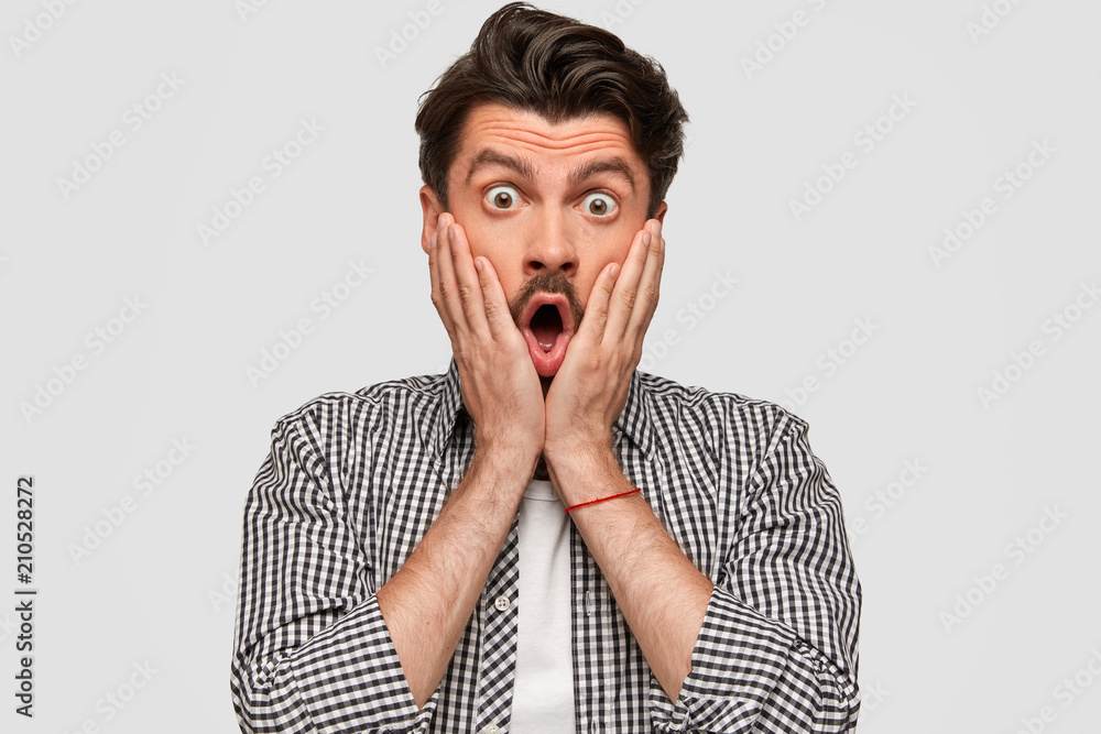 It`s amazing! Attractive young male hipster with bated breath, keeps hands on cheeks, has eyes popped out, wears fashionable clothes, isolated over white background. People and reaction concept It`s amazing! Attractive young male hipster with bated breath, keeps hands on cheeks, has eyes popped out, wears fashionable clothes, isolated over white background. People and reaction concept