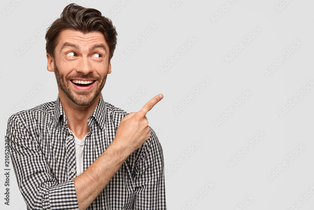 Glad satisfied male in stylish shirt, indicates with index finger at upper right corner, has joyful expression, suggests to use this blank space for advertising content, isolated on white background Glad satisfied male in stylish shirt, indicates with index finger at upper right corner, has joyful expression, suggests to use this blank space for advertising content, isolated on white background