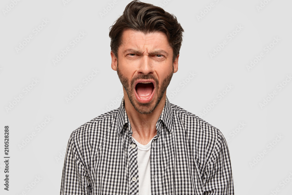 Angry unshaven male student screams with annoynace, frowns face, has quarrel with groupmates, dressed in stylish checkered shirt, isolated over white background. Negative human emotions concept