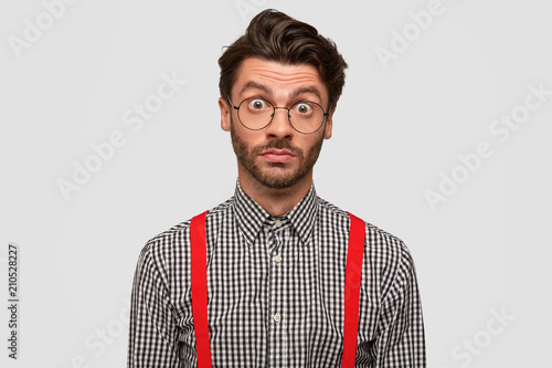 Photo of suprised male student with trnedy hairstyle, dressed in checkered shirt, recieves information about coming session and has many exams, isolated over white background. Stupefied man indoor photo