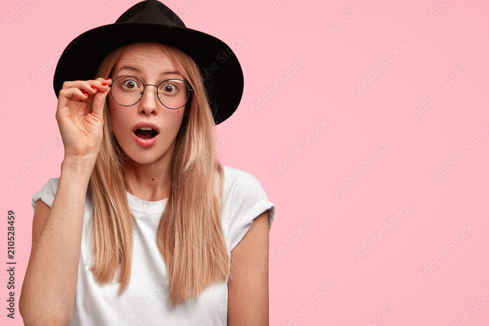 Astonished lovely young female designer reacts on unexpected task and much work, keeps hand on rim of spectacles, wears big stylish black hat, isolated over pink background, expresses surprisement Astonished lovely young female designer reacts on unexpected task and much work, keeps hand on rim of spectacles, wears big stylish black hat, isolated over pink background, expresses surprisement