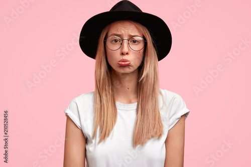 Unsatisfied blonde young female purses lower lip, looks with miserable upset expression, being offended by close person, dressed casually, isolated over pink background. Negative feeling concept