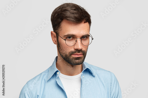 Portrait of serious young male journalist wears round glasses and blue shirt, has confident look at camera, recieves important task from boss, listens attentively, isolated over white background. © Wayhome Studio