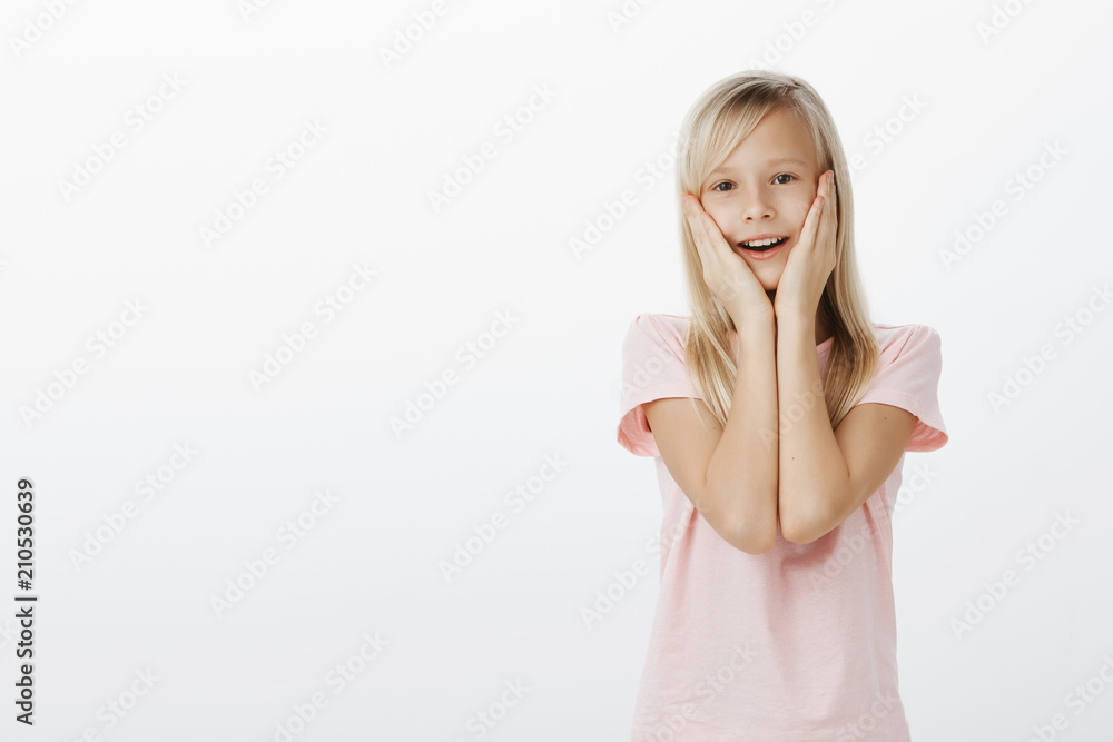 Little girl touched seeing cute pony. Indoor shot of pleased adorable female child with blond hair, holding hands on cheeks and smiling, being surprised and happy seeing something great over gray wall Little girl touched seeing cute pony. Indoor shot of pleased adorable female child with blond hair, holding hands on cheeks and smiling, being surprised and happy seeing something great over gray wall