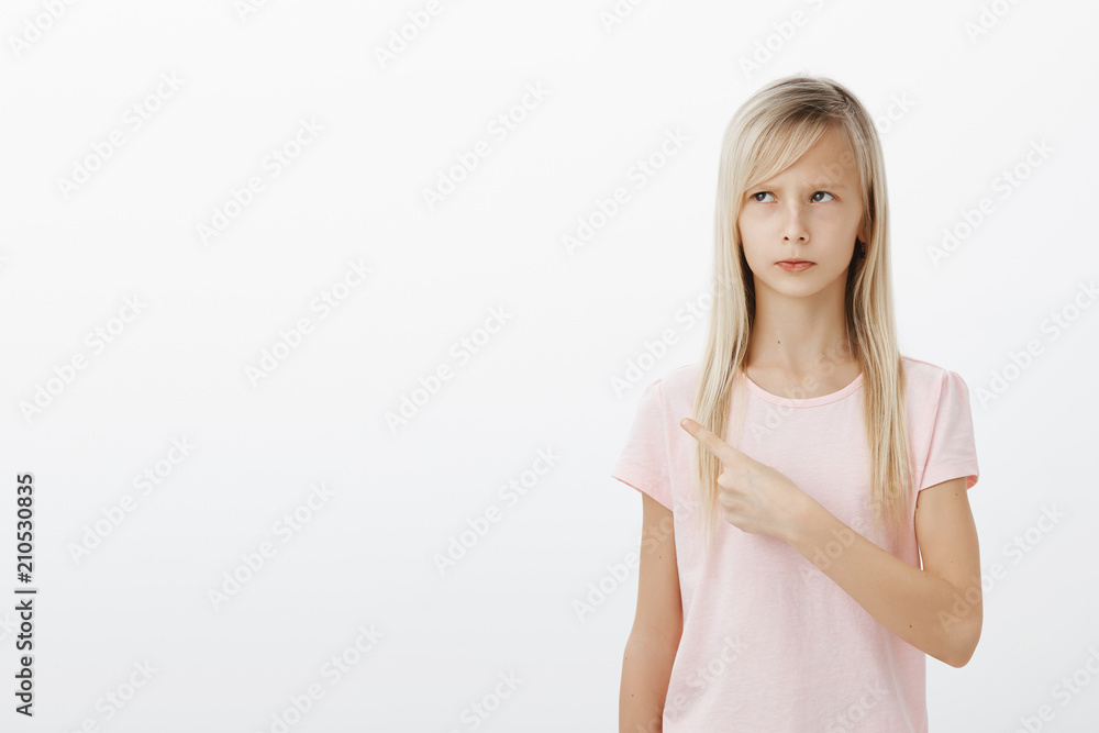 Girl showing mom where she hidden sweets in room. Portrait of offended upset cute child with blond hair, frowning, feeling sad and gloomy, pointing and looking at upper left corner over gray wall Girl showing mom where she hidden sweets in room. Portrait of offended upset cute child with blond hair, frowning, feeling sad and gloomy, pointing and looking at upper left corner over gray wall