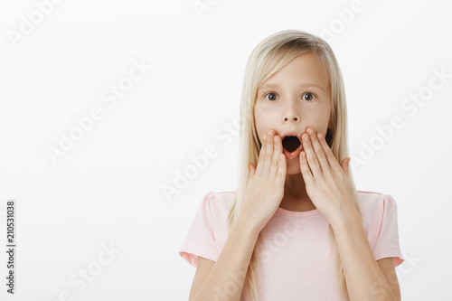 Wow, kid shocked hearing amazing news about favorite cartoon. Indoor shot of amazed and surprised happy blond daughter, gasping, standing with hands near opened mouth and popping eyes, being stunned
