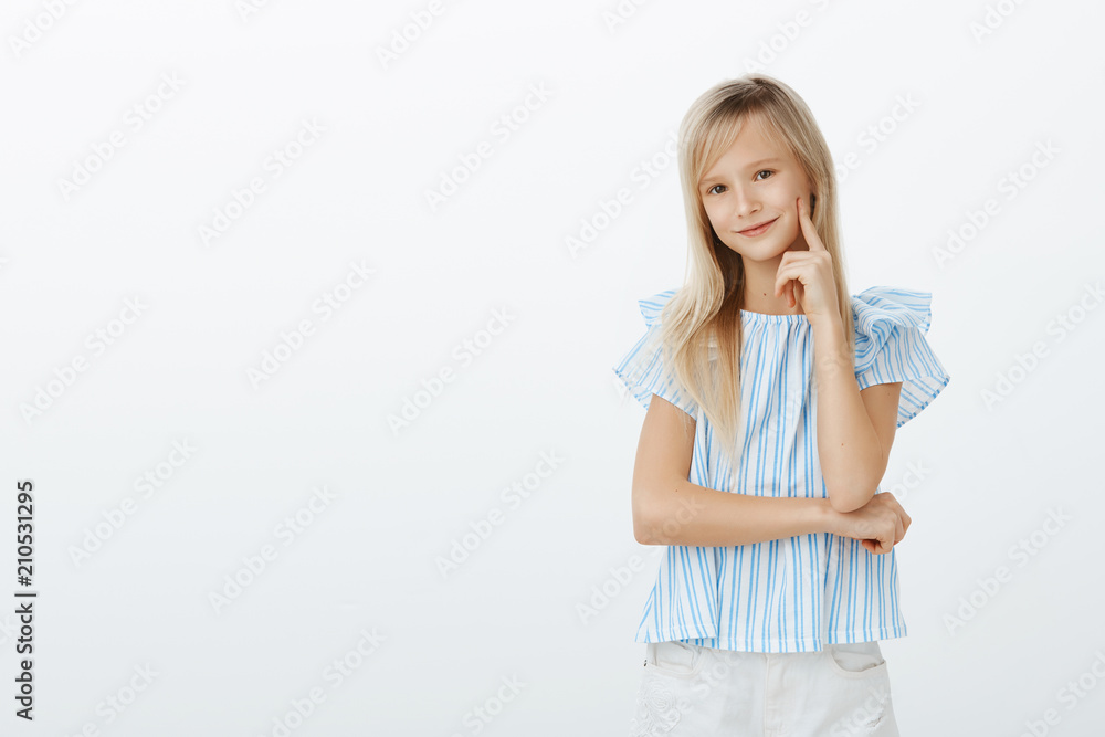 Little fashionable girl knows everything about cute dresses. Pleased happy emotive child with blond hair, holding finger on cheek and smiling broadly, having great idea in mind over gray wall Little fashionable girl knows everything about cute dresses. Pleased happy emotive child with blond hair, holding finger on cheek and smiling broadly, having great idea in mind over gray wall