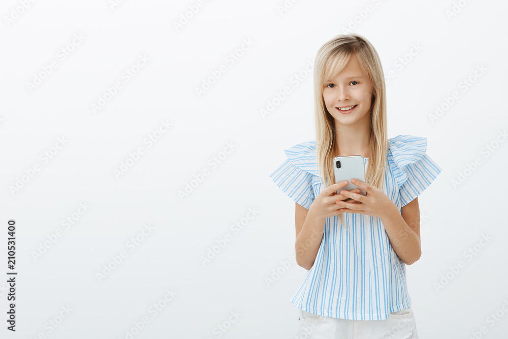 Little kid stole dads cellphone to play games. Portrait of charming happy young girl with blond hair, holding smartphone and smiling broadly, watching cartoons or messaging with friends over gray wall Little kid stole dads cellphone to play games. Portrait of charming happy young girl with blond hair, holding smartphone and smiling broadly, watching cartoons or messaging with friends over gray wall