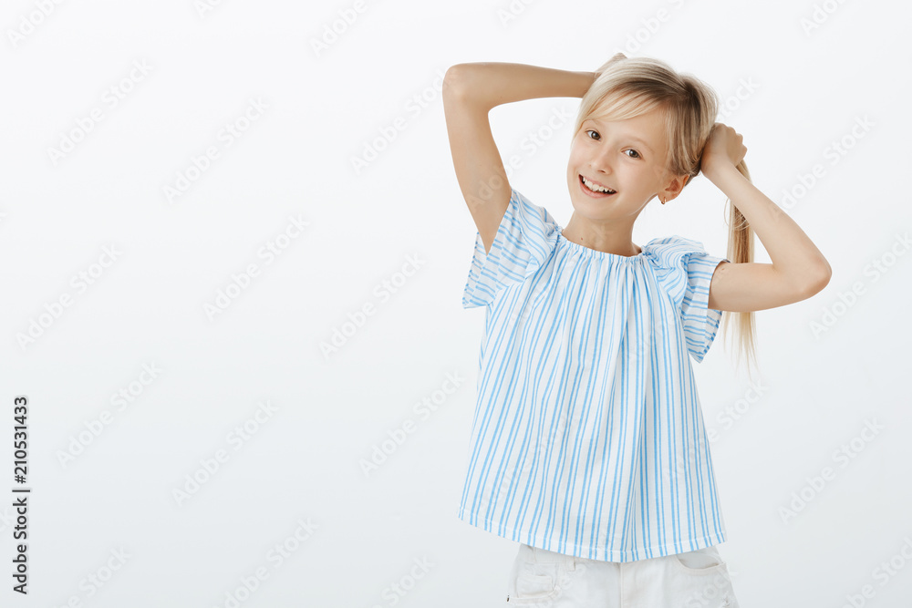 Little fashionable girl showing her first earrings to friend. Portrait of happy playful european child, holding fair hair in hands, making braids and smiling broadly, fooling around over gray wall Little fashionable girl showing her first earrings to friend. Portrait of happy playful european child, holding fair hair in hands, making braids and smiling broadly, fooling around over gray wall