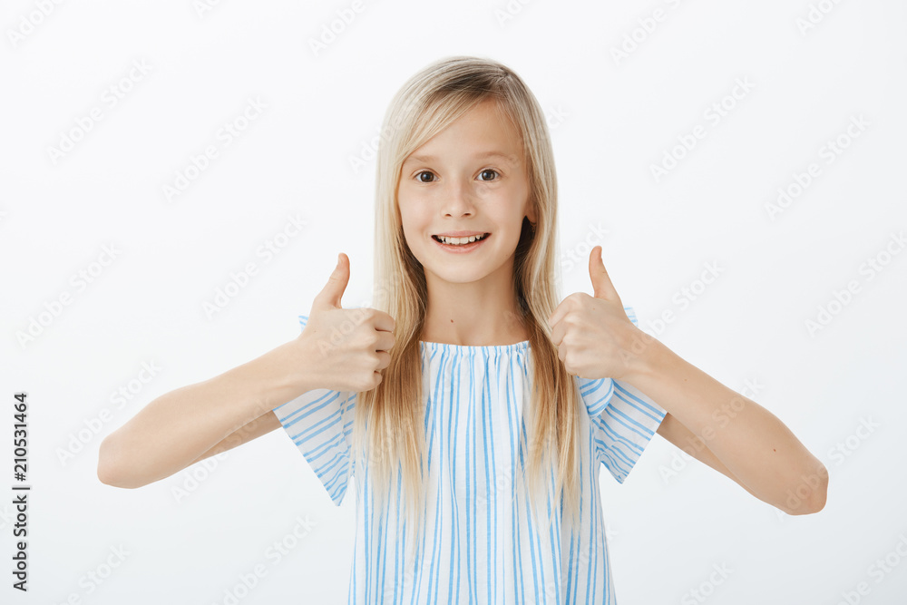 Supportive little friend wants to cheer mates. Happy positive cute girl with blond hair, raising thumbs up and giving approval, smiling broadly while liking great idea, standing over gray wall Supportive little friend wants to cheer mates. Happy positive cute girl with blond hair, raising thumbs up and giving approval, smiling broadly while liking great idea, standing over gray wall
