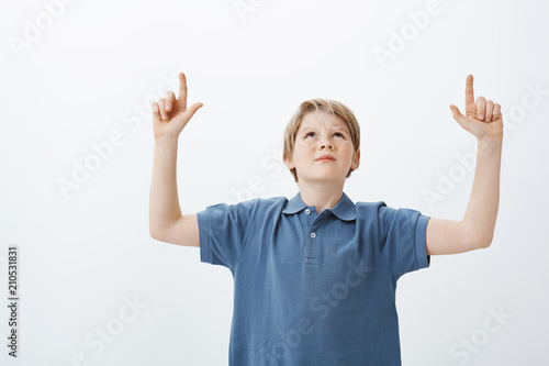 Indoor shot of displeased doubtful man with blond hair, looking and pointing up with raised index fingers, frowning, being unsure and annoyed with loud neighbours, trying to study over grey wall