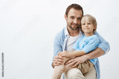 Studio shot of happy positive father and son with vitiligo, hugging and smiling broadly at camera, being pleased and satisfied spending time together, feeling love and caring emotions to family