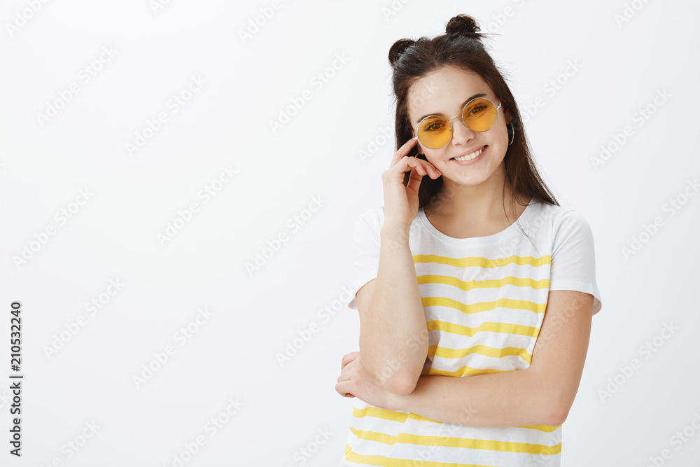 Waist-up shot of carefree cute and fashionable caucasian female model in trendy sunglasses and striped yellow t-shirt, touching rim of glasses and smiling broadly, listening with interest story Waist-up shot of carefree cute and fashionable caucasian female model in trendy sunglasses and striped yellow t-shirt, touching rim of glasses and smiling broadly, listening with interest story