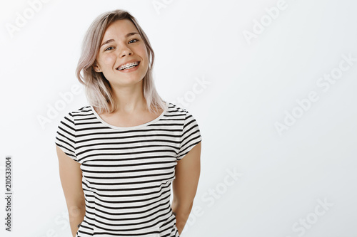 Studio shot of cute caucasian girlfriend with blond hair and braces on teeth standing in casual striped t-shirt and smiling broadly at camera, holding hands behind, enjoying great sunny day photo