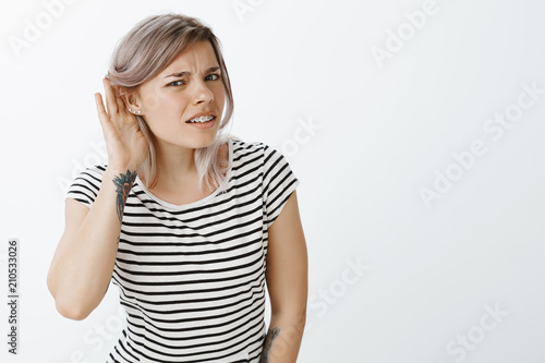 Repeat louder  cannot hear your whisper. Portrait of intense confused attractive female student in striped t-shirt  bending towards camera  holding hand near ear  gossiping or eavesdropping
