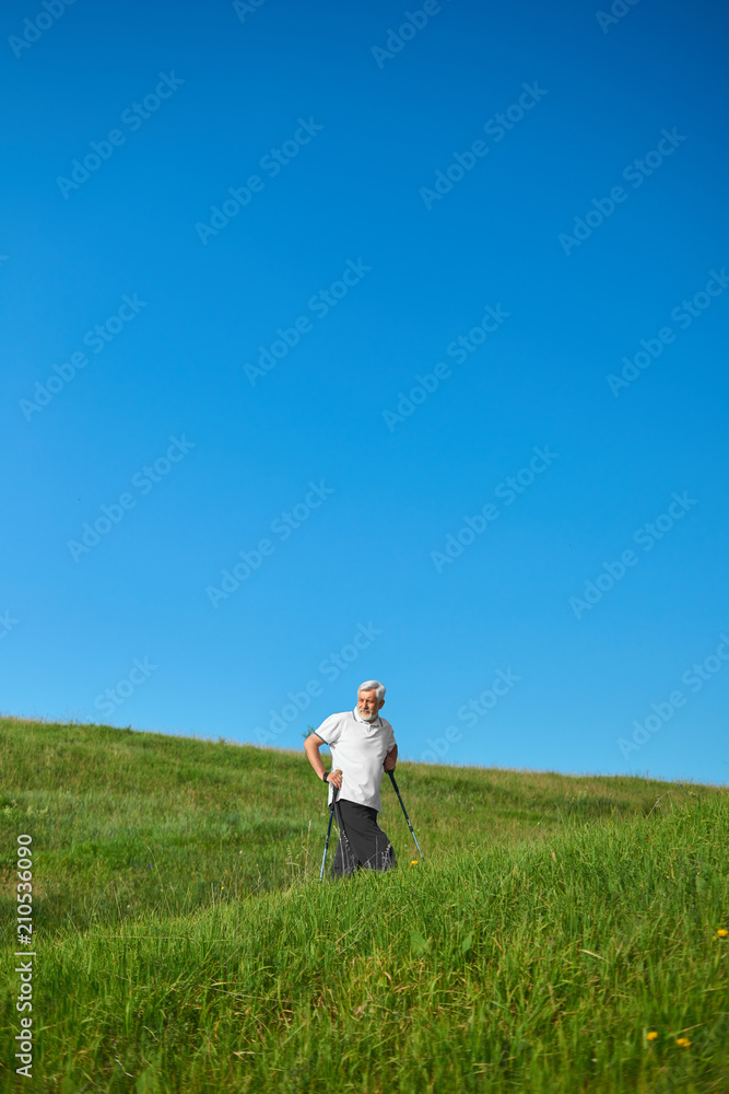 Old man walking on hills with tracking sticks. Walking on green field. Summer day, good weather, outdoor sport. Healthy lifestle. Male model wearing white polo shirt and black trousers. Old man walking on hills with tracking sticks. Walking on green field. Summer day, good weather, outdoor sport. Healthy lifestle. Male model wearing white polo shirt and black trousers.