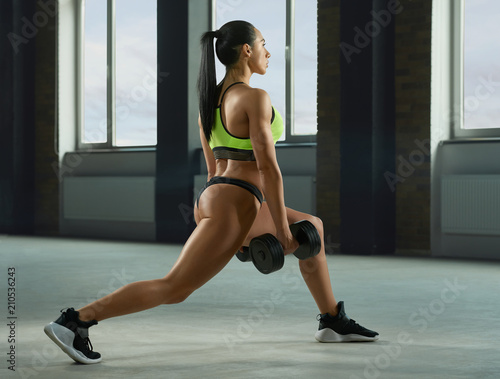 Sideview of athletic girl performing fallouts keeping dumbbells in spacy gym with panoramic windows. Having strong, fit body with heatlthy tanned skin, muscles. Doing fitness exercises.