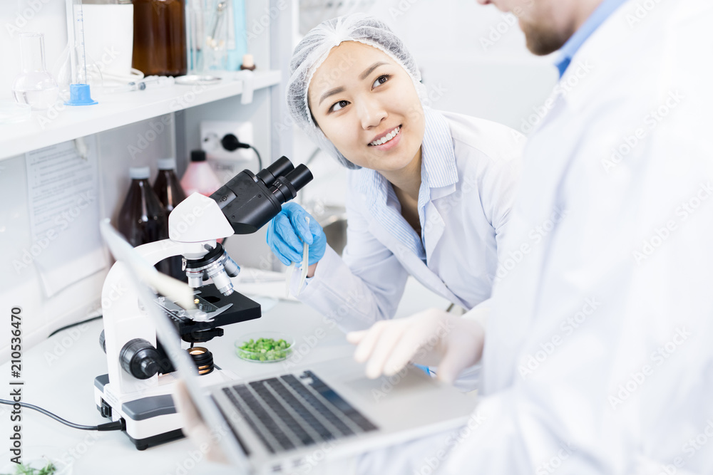 Crop view of smiling female microbiologist standing at eyepiece of microscope with samples of green vegetables looking at colleague typing results of analysis on laptop Crop view of smiling female microbiologist standing at eyepiece of microscope with samples of green vegetables looking at colleague typing results of analysis on laptop