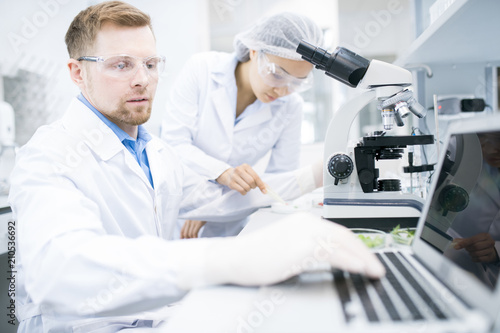 Side view of male microbiologist in white laboratory coat typing on laptop keyboard data received during testing of food nutrition properties under microscope with female assistant on background.
