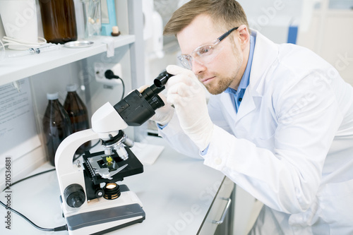 Crop view of man in glasses and laboratory coat standing at desk and looking at eyepiece of microscope with green vegetable sample lying on it