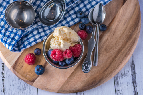 A scoop of vanilla ice cream with raspberries and blueberries on a wooden board and blue and white dish towel.
