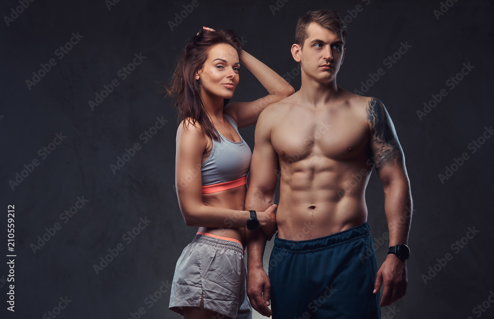 Attractive couple, a slim brunette female wearing sports bra and shorts and handsome shirtless guy cuddling in a studio on a dark textured background.