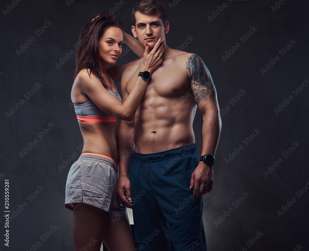 Attractive couple, a slim brunette female wearing sports bra and shorts and  handsome shirtless guy cuddling in a studio on a dark textured background.  Stock Photo