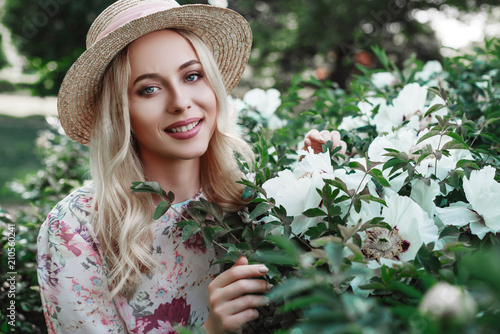 Outdoor close up portrait of beautiful happy smiling woman wearing straw boater hat, dress posing in the blooming garden