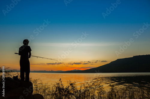 Silhouette a man standing fishing with empty water surface of lake landscape view with mountain background at sunset time