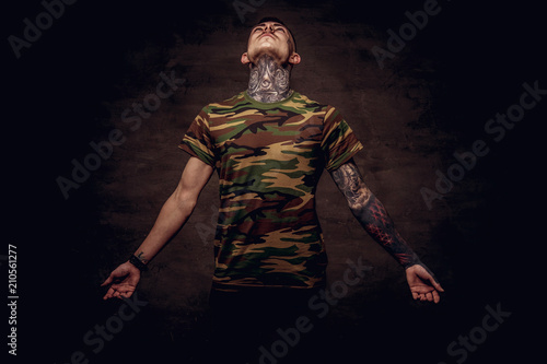 Tattoed young guy in a military t-shirt pose at the studio. Isolated on dark textured background.