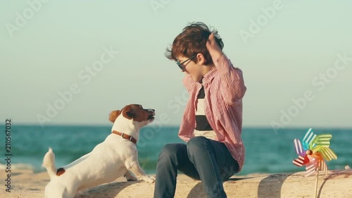 Slow motion a happy boy sits on a log on the seashore and plays emotionally with his dog breed Jack Russell, holds a cookie in his hand and feeds a pet, teasing him with a treat, dog jumps, windmill