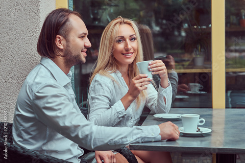 Attractive couple - charming blonde female dressed in a white blouse and bearded male with a stylish haircut dressed in a white shirt during a date at cafe outdoors.