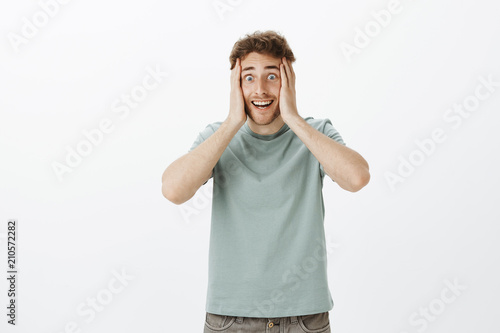 Portrait of excited crazy european guy in t-shirt, holding hands on face and smiling with weird funny expression, being happy and overwhelmed with positive emotions hearing great news