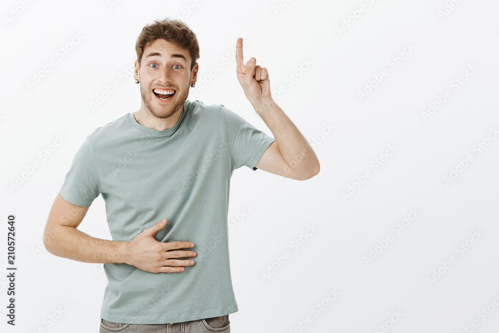Portrait of friendly good-looking Caucasian man with bristle and fair hair, raising index finger and pointing up, laughing out loud and holding hand on chest while having fun over gray background Portrait of friendly good-looking Caucasian man with bristle and fair hair, raising index finger and pointing up, laughing out loud and holding hand on chest while having fun over gray background
