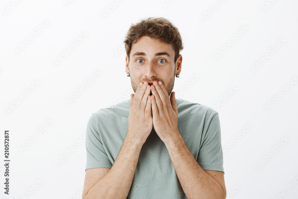 Portrait of happy european male model in earrings, smiling and covering mouth with palms, being amazed and pleased with great news, standing satisfied while hearing surprising revelation Portrait of happy european male model in earrings, smiling and covering mouth with palms, being amazed and pleased with great news, standing satisfied while hearing surprising revelation
