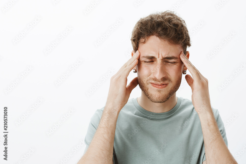Close-up shot of intense caucasian fair-haired guy suffering from pain, frowning and closing eyes, touching temples with palms, trying focus but feeling migraine or headache, standing over grey wall Close-up shot of intense caucasian fair-haired guy suffering from pain, frowning and closing eyes, touching temples with palms, trying focus but feeling migraine or headache, standing over grey wall