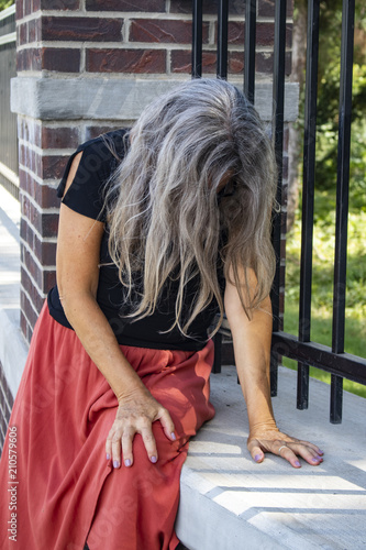 Woman in dress sits by fence with her head bowed and face covered with long gray hair sick or sad or in distress