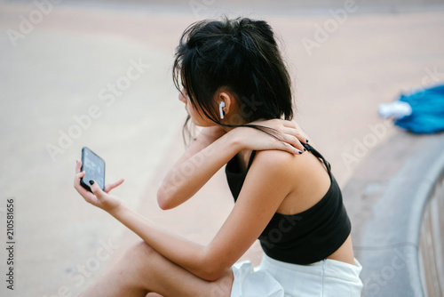 A young and pretty Chinese Asian teenager sits next to her skateboard in a skate park during the day. She is listening to music on her smartphone through her wireless headset.