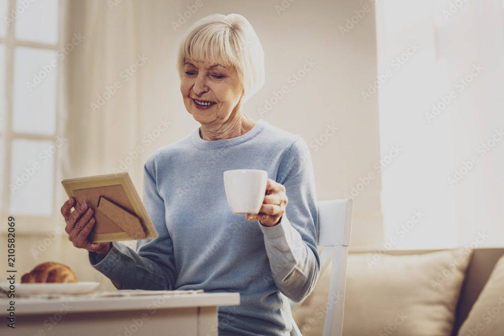 Pleasant memories. Joyful elderly woman looking at the photo while having a cup of coffee Pleasant memories. Joyful elderly woman looking at the photo while having a cup of coffee