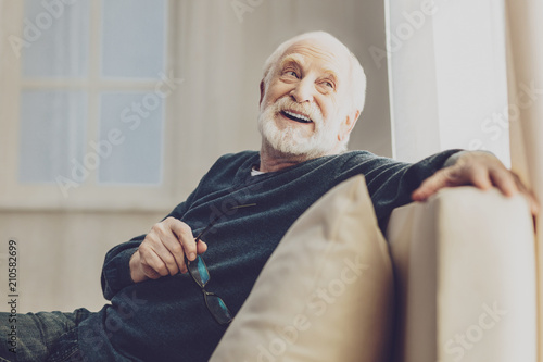 Pleasurable time. Joyful senior man smiling while enjoying his rest at home