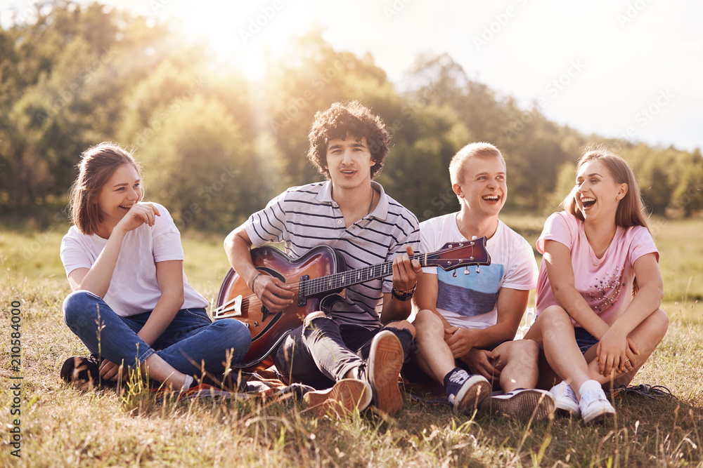 Fototapeta premium Happy female and male students enjoy picnic outdoor, sit grouped together, laugh and joke among themselves, sing songs to guitar, admire beautiful nature. People, lifestyle, amuzement concept