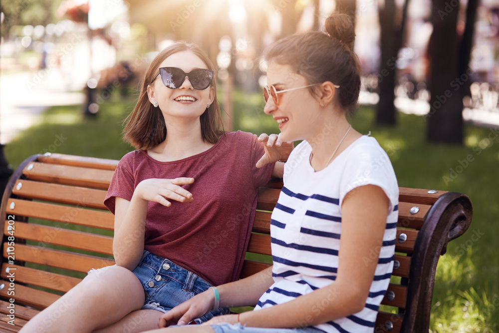 Funny female teenagers laugh joyfully while tell each other funny stories, dressed in casual clothes, sit on wooden bench, being in good mood, express positive emotions. Smiling women friends Stock Photo | Funny female teenagers laugh joyfully while tell each other funny stories, dressed in casual clothes, sit on wooden bench, being in good mood, express positive emotions. Smiling women friends Stock Photo |