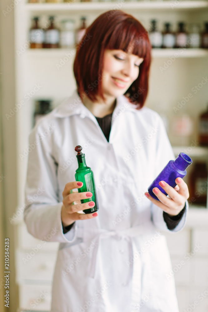 Attractive young lady pharmacist with a lovely smile standing working and holding glass vintage bottles of medicines in the pharmacy Attractive young lady pharmacist with a lovely smile standing working and holding glass vintage bottles of medicines in the pharmacy