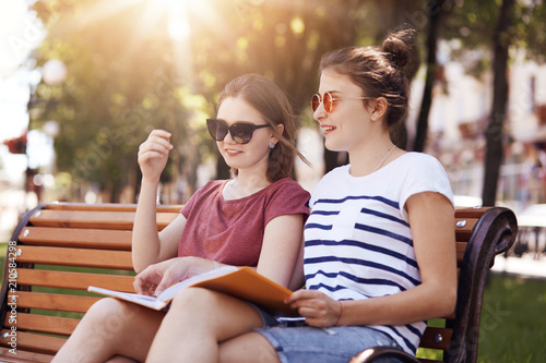 Outside shot of cheerful female friends, have gentle smiles, prepare for final exam together, sit on bench during hot summer day, being satisfied with something. People and lifestyle concept