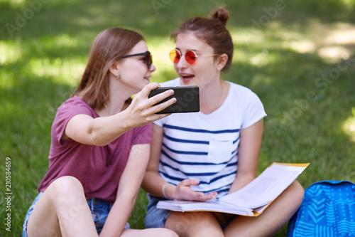 Youth and technology concept. Positive young best female friends sit closely  read book and pose for making selfie portrait  focus on modern smart phone  pose against green nature background