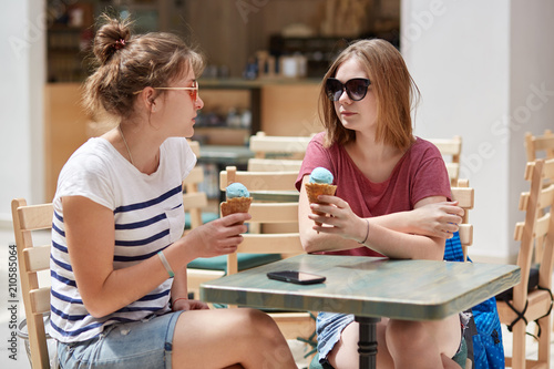 Friends, recreation and summer concept. Two beautiful female teenagers eat cold delicious ice cream, talk with each other, discuss paln for going to sea, enjoy pleasant friendly conversation