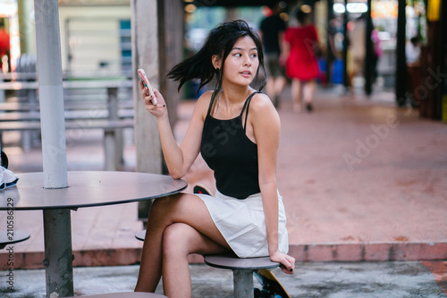 An image of a pretty  sporty  attractive  and too-cool-for-school Chinese Asian teenager at a hawker center looking away as she holds her smartphone.