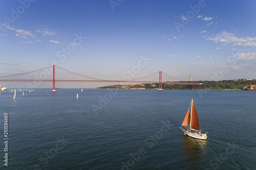 Aerial view of a beautiful sail boat on the Tagus River with the 25 of April Bridge on the background, in the city of Lisbon, Portugal © Tiago Fernandez
